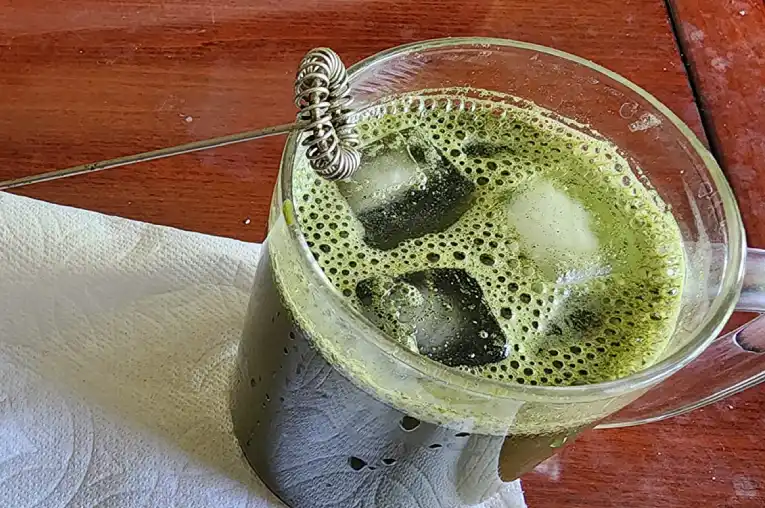 a cup of organifi green juice powder mix with water with ice cubes on a wooden table.